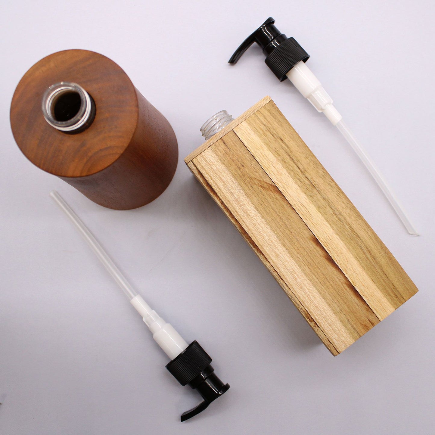 Dos dispensadores de jabón de madera natural: uno cilíndrico oscuro y otro cuadrado claro, con dos bombas negras.