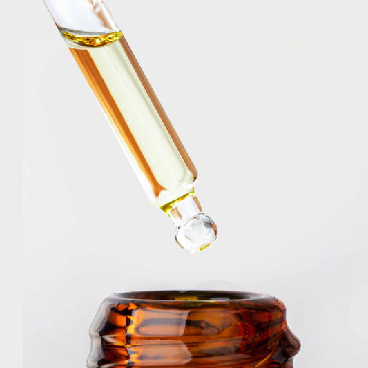 Gotero de vidrio con serum facial dorado de Vitamina C de Ancient Wisdom cayendo sobre frasco ámbar. Fondo blanco.