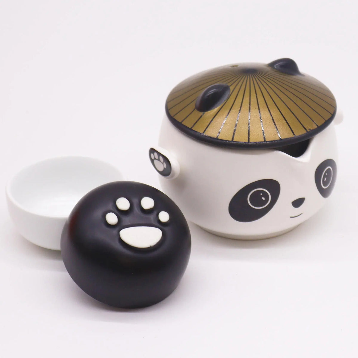 Tetera de cerámica blanca con diseño de panda, tapa dorada tipo sombrero, dos tazas pequeñas y huella de pata negra