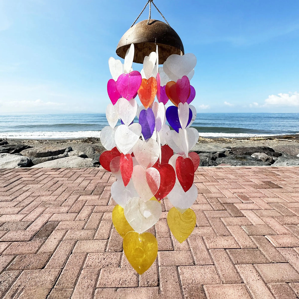 Campanilla de viento con base de coco y corazones de nácar capiz de colores colgando ante un paisaje de playa.