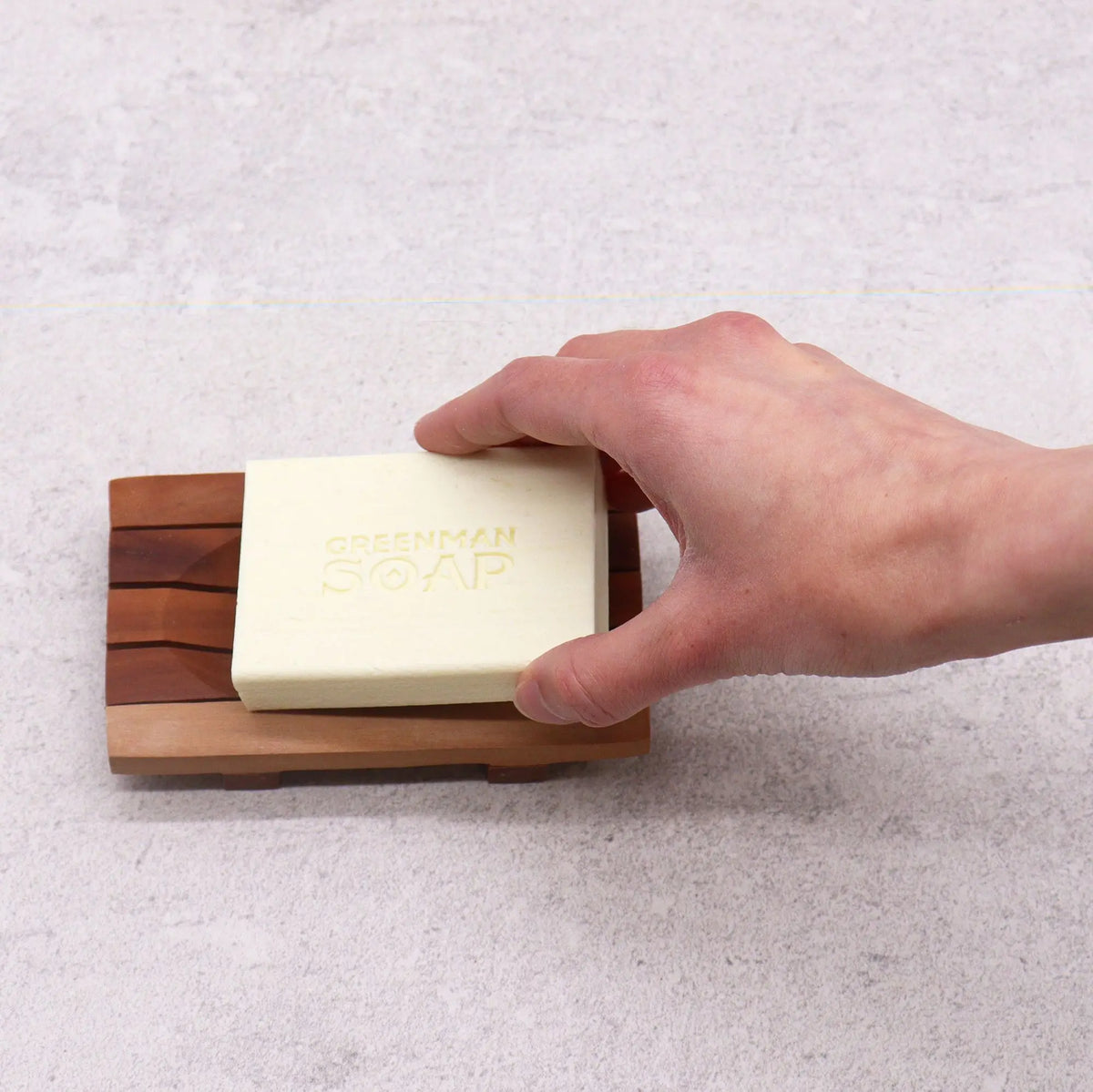 Jabonera rectangular de caoba con rejilla drenante y una pastilla de jabón beige, sostenida por una mano sobre fondo gris.