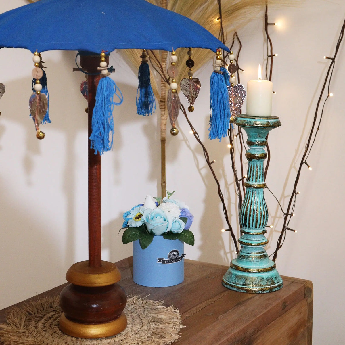 Sombrilla artesanal de Bali color turquesa con borlas y abalorios, sobre base de madera, junto a un candelabro rústico.