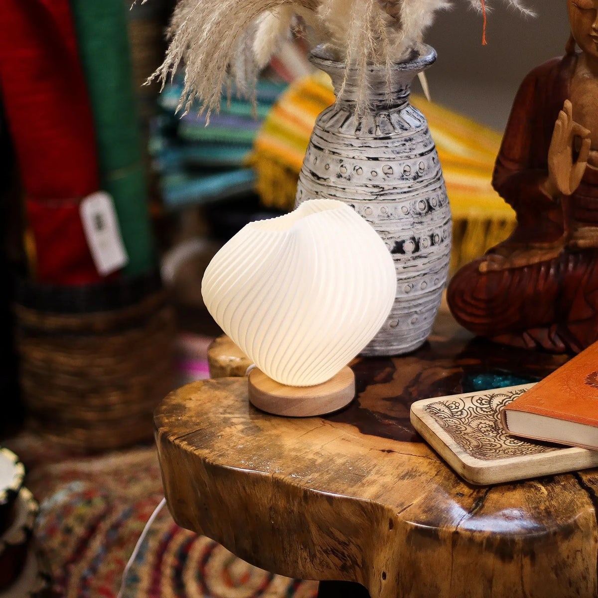 Lámpara de mesa blanca con forma de concha espiral sobre base de madera, emitiendo luz cálida en una mesa rústica.