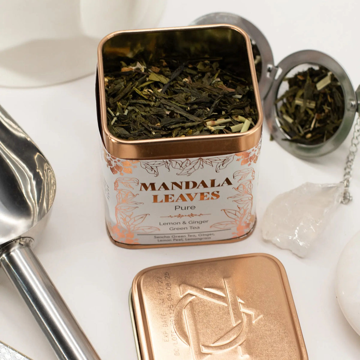 Lata cuadrada color cobre con té verde, jengibre y limón, junto a una cuchara metálica y un infusor sobre fondo blanco.