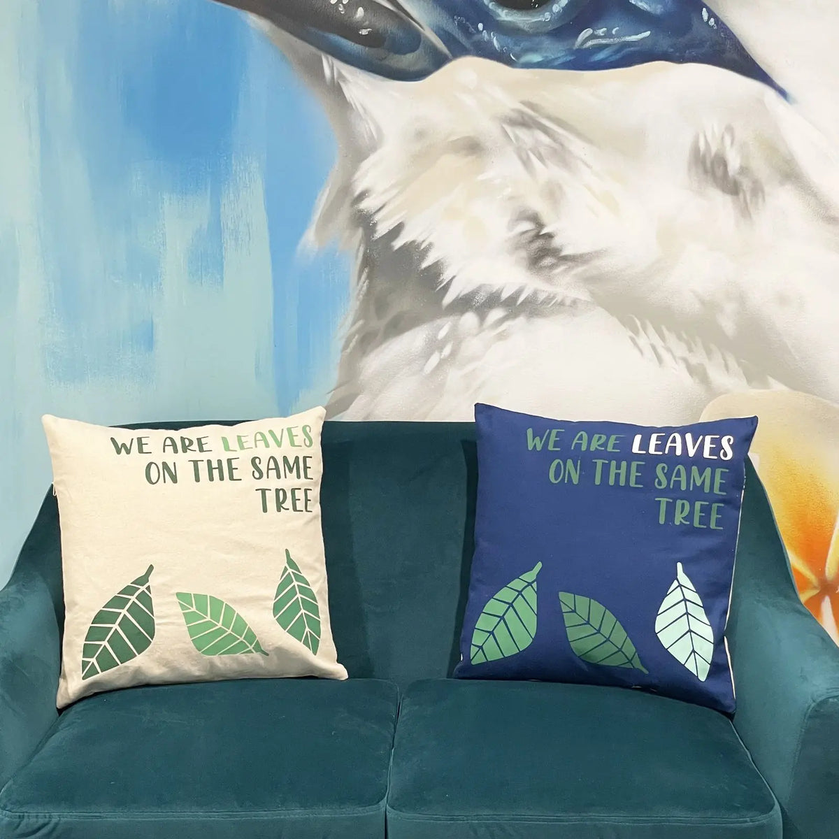 Dos fundas de cojín de algodón con estampado de hojas y texto sobre sofá verde, una en color natural y otra en azul.