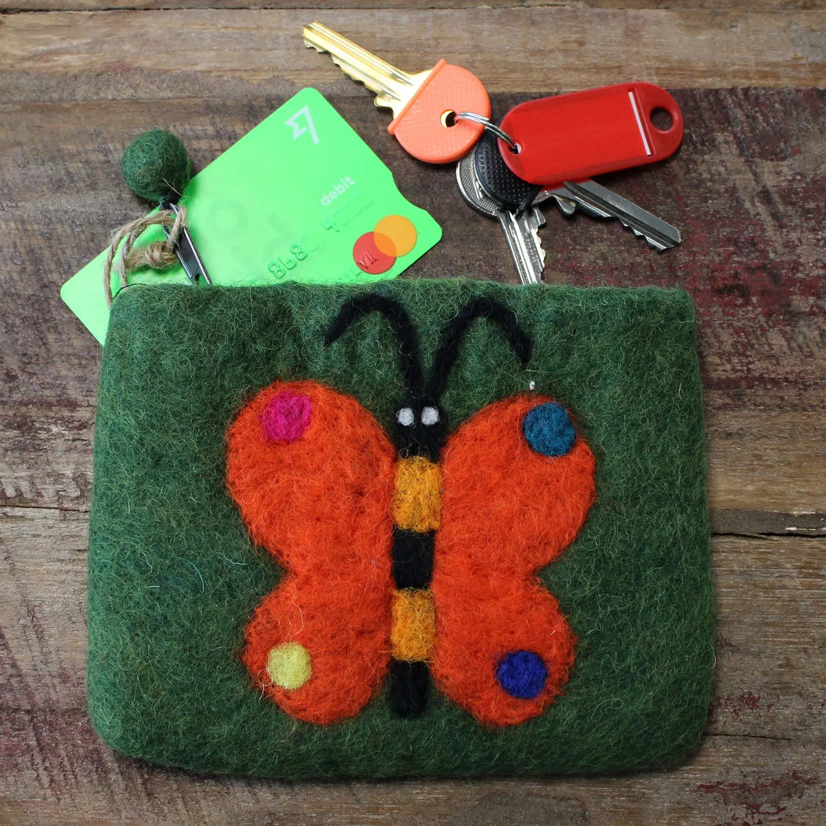 Estuche de fieltro verde con una mariposa naranja aplicada, sobre una superficie de madera con llaves y tarjeta dentro.