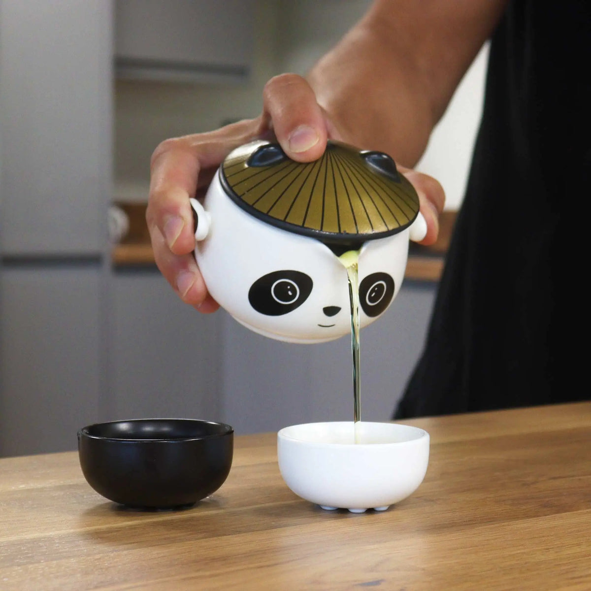Tetera de cerámica blanca con diseño de panda y tapa de sombrero vertiendo té en una taza sobre mesa de madera.