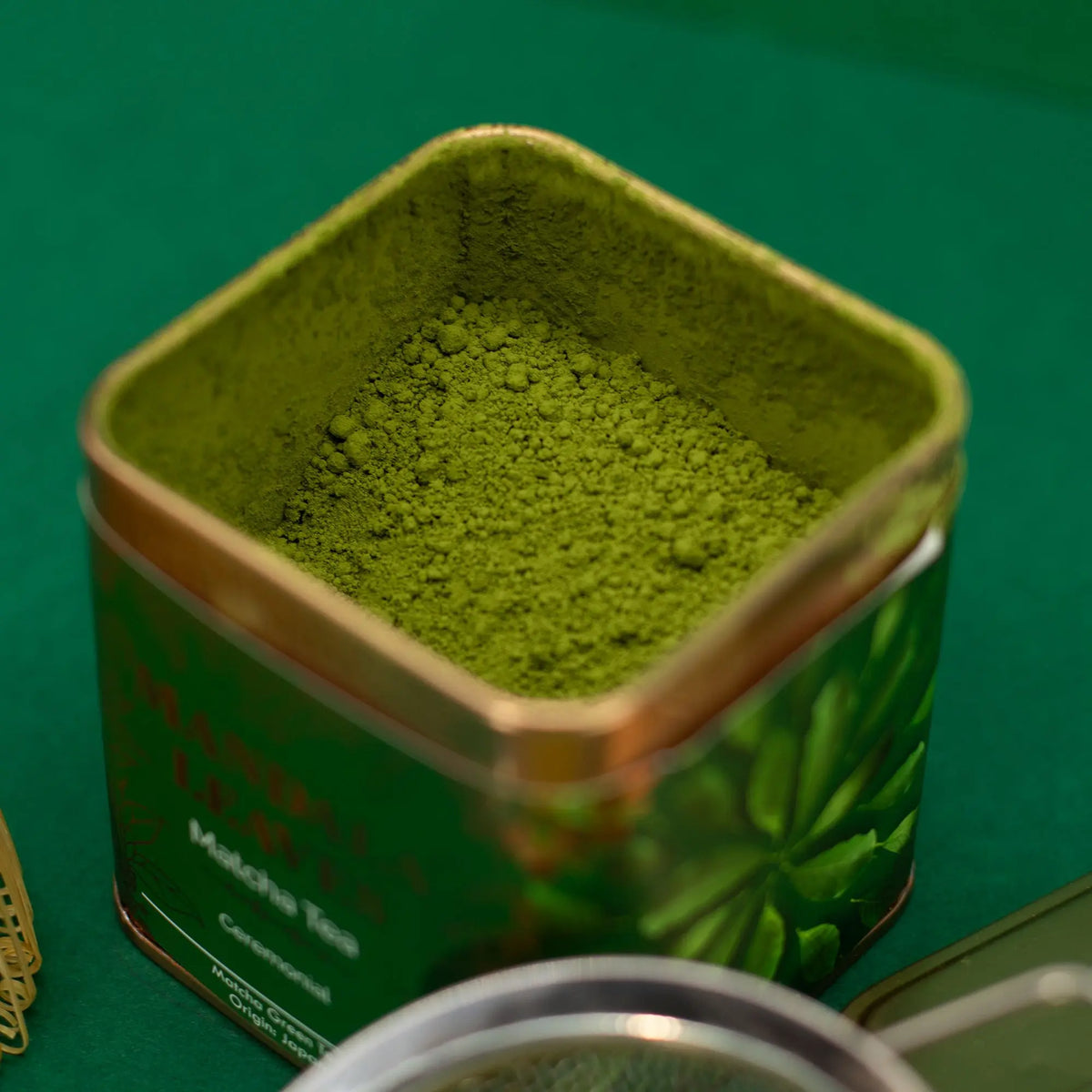 Lata cuadrada dorada abierta llena de polvo de té matcha verde intenso sobre una superficie del mismo tono.