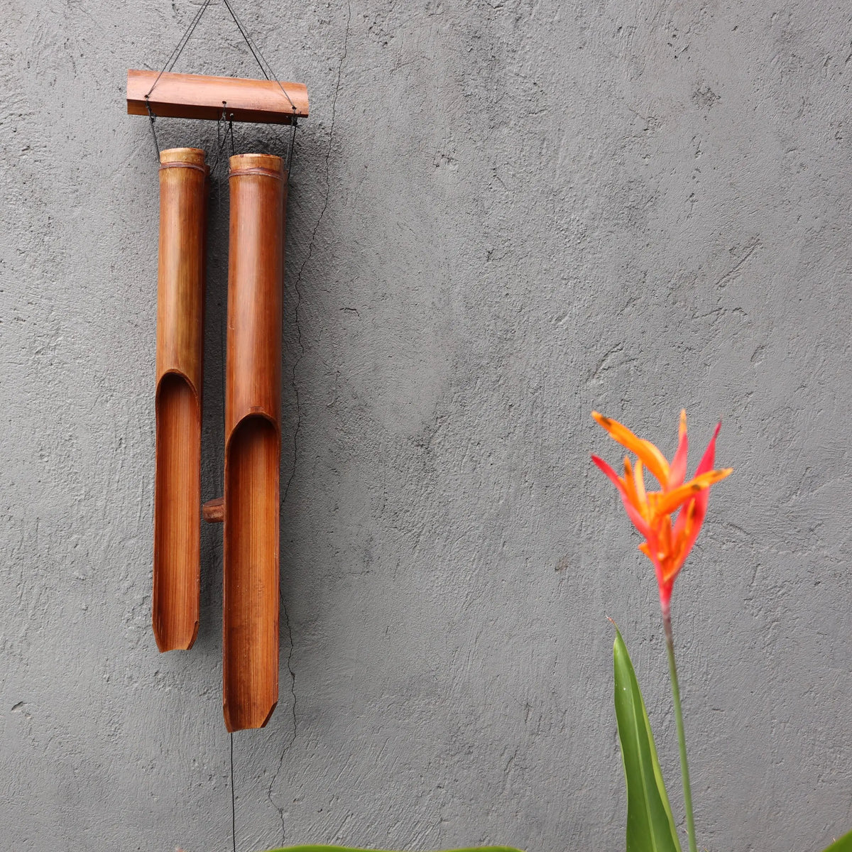 Campanilla de viento de bambú con dos tubos largos, acabado natural, colgada frente a una pared gris y una flor roja.