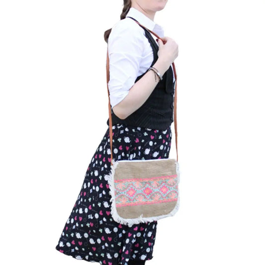Bolso bandolera de yute beige con flecos blancos y bordado geométrico en tonos rosa y azul, usado sobre el hombro.