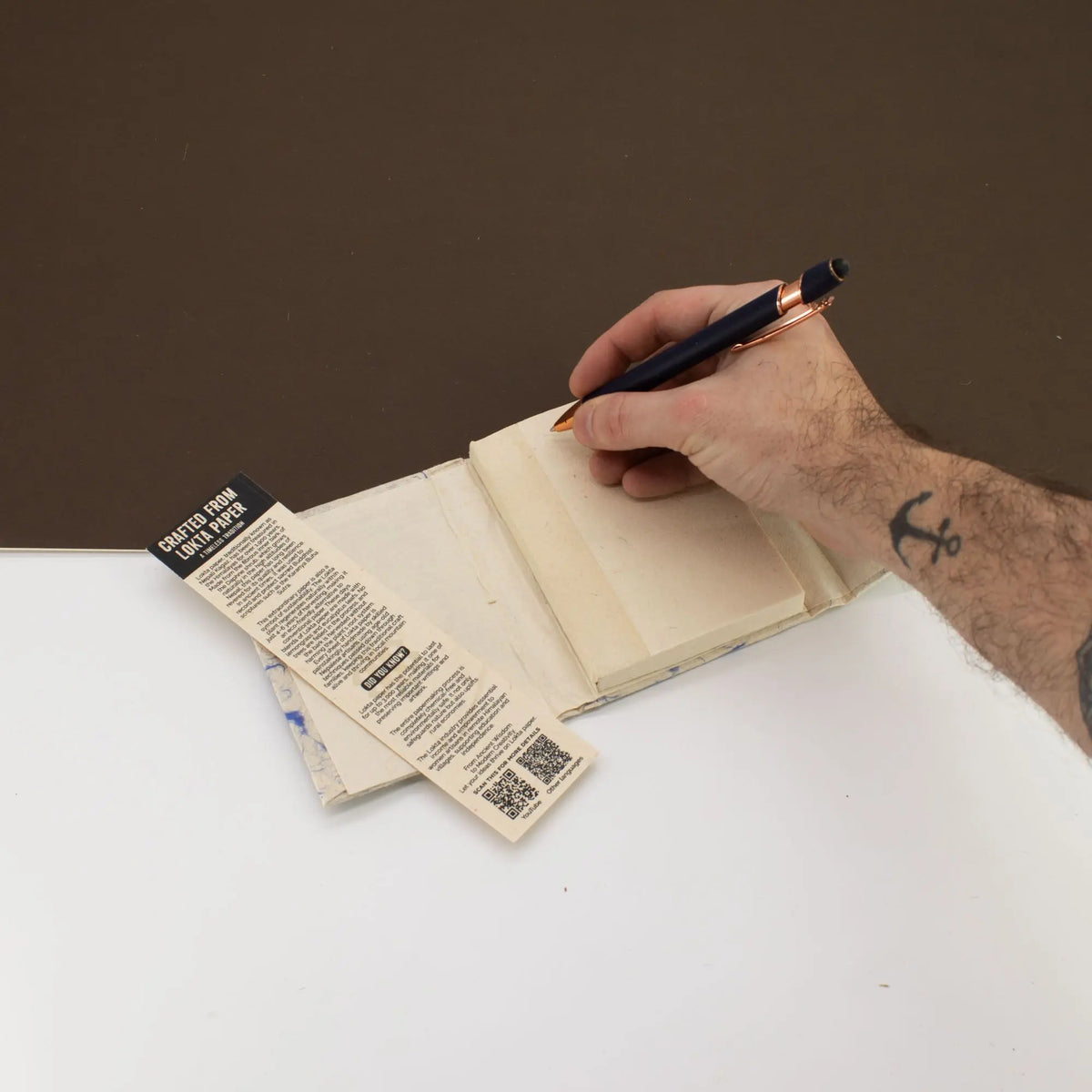 Cuaderno blanco con cubierta kraft, una mano escribiendo con un bolígrafo negro y plateado.