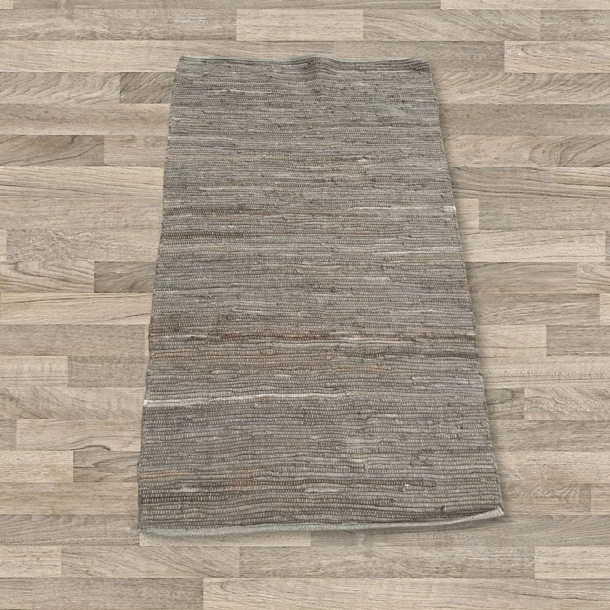 Alfombra pequeña rectangular de algodón color carbón, tejida a mano con textura rústica sobre suelo de madera clara.