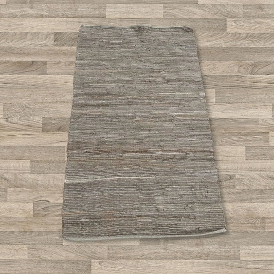 Alfombra pequeña rectangular de algodón color carbón, tejida a mano con textura rústica sobre suelo de madera clara.