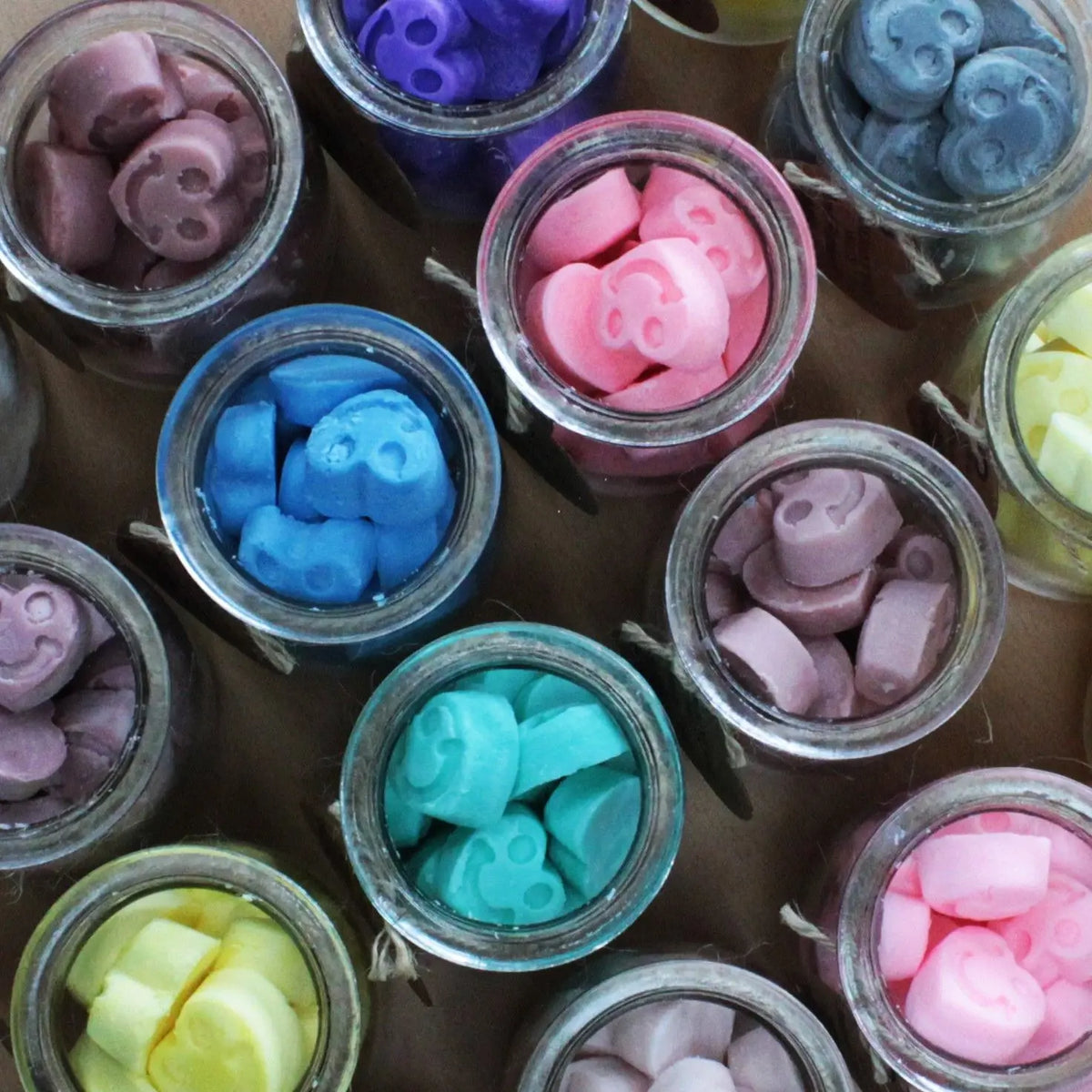 Frascos de vidrio con pequeñas figuras de cera de soja en forma de caritas felices de diversos colores.