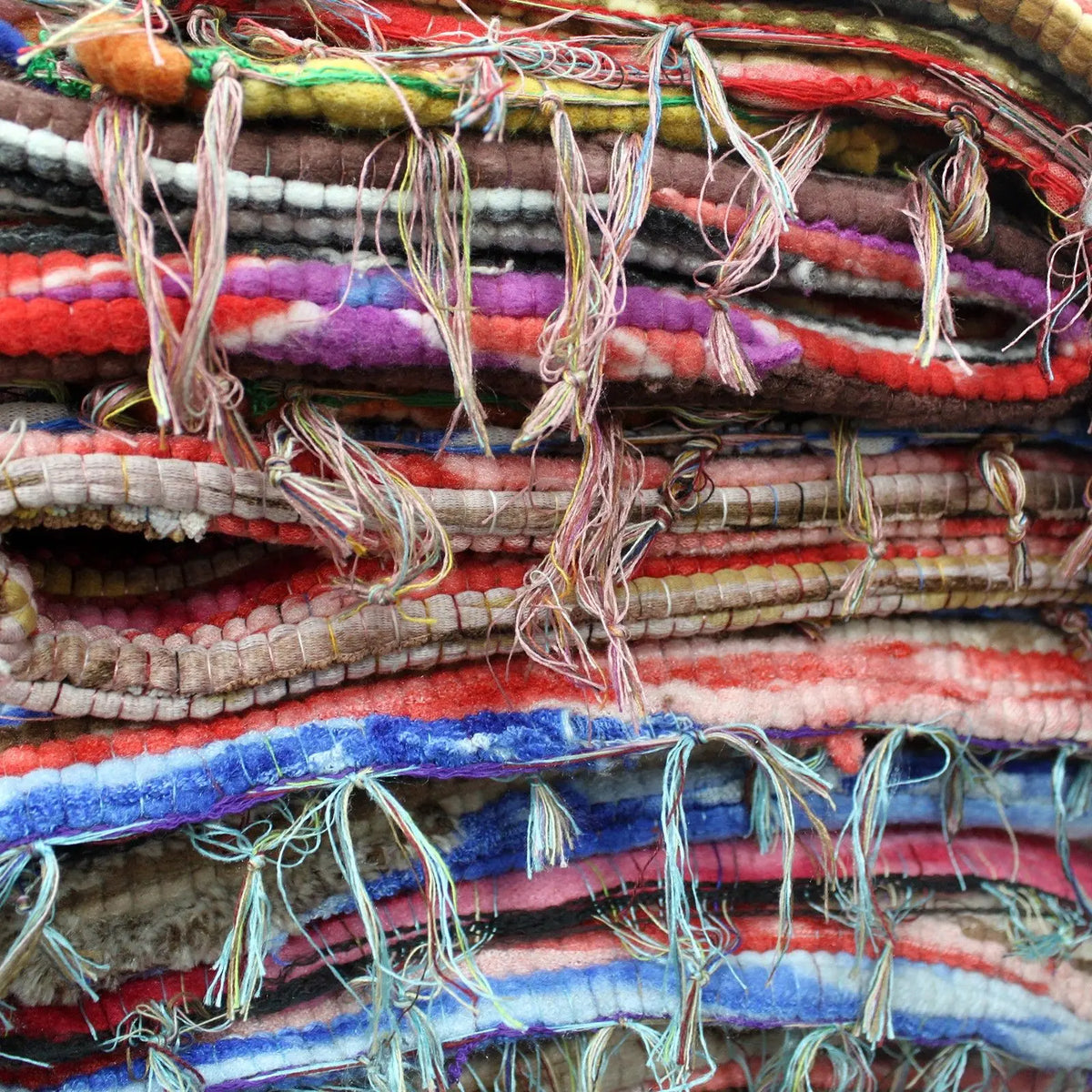 Pila de alfombras artesanales de trapo multicolor con textura de tejido grueso y flecos deshilachados en los bordes.