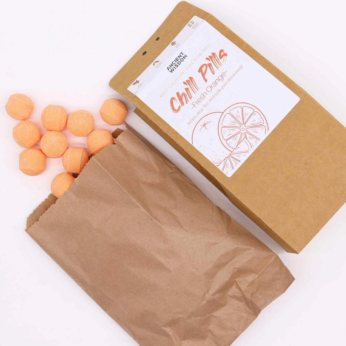 Mini bombas de baño efervescentes color naranja saliendo de una bolsa de papel kraft sobre fondo blanco.
