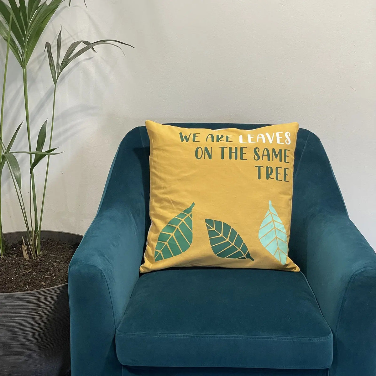 Funda de cojín de algodón amarillo con estampado de hojas y el texto "We are leaves on the same tree" en sillón azul.