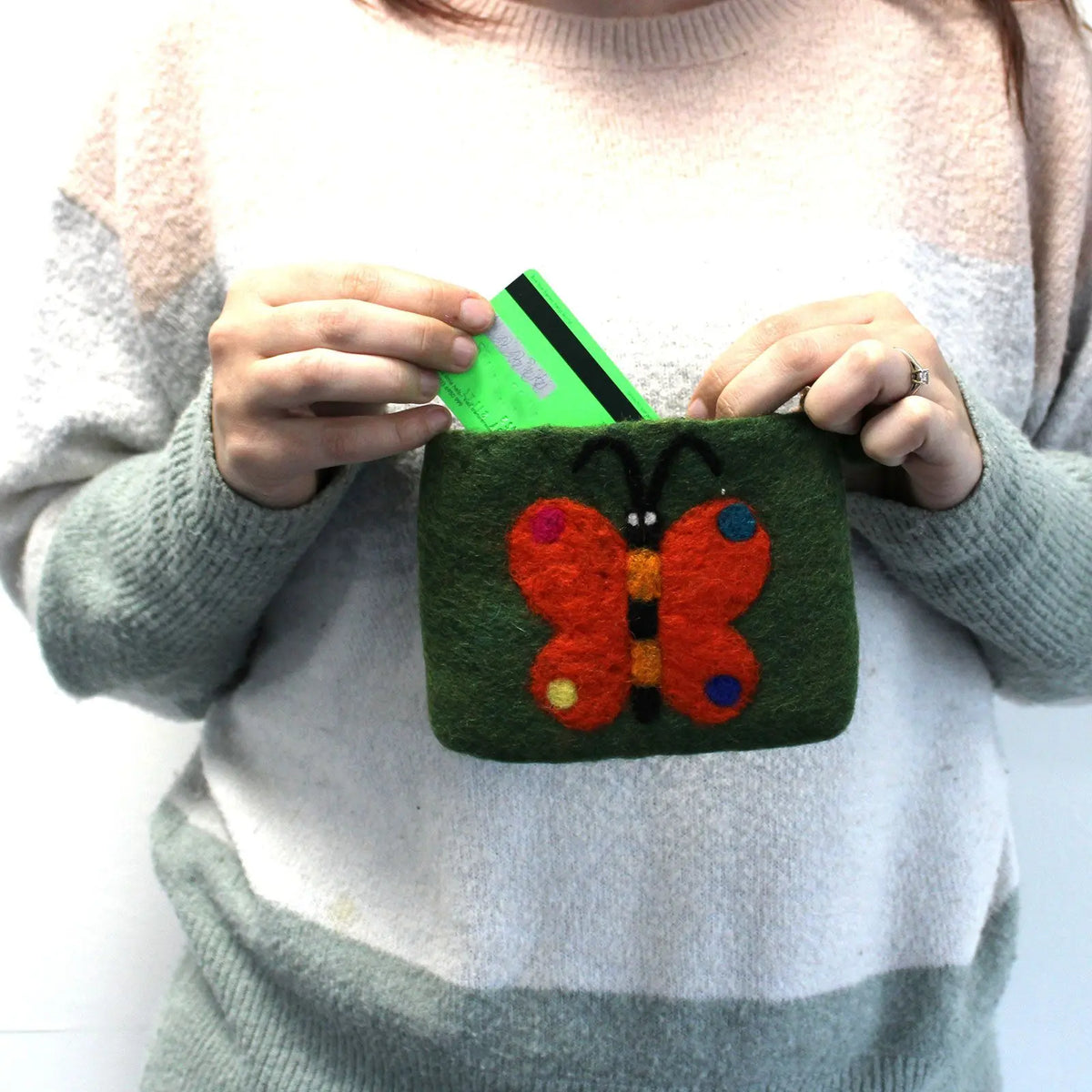Estuche de fieltro verde con una mariposa naranja de fieltro, sostenido por manos que introducen una tarjeta verde.