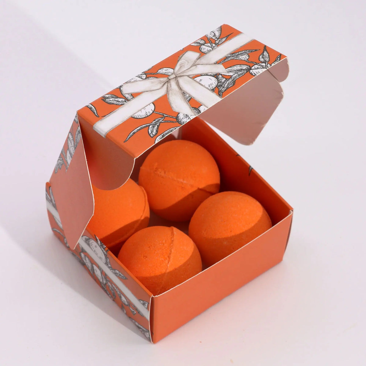Caja naranja con cuatro bombas de baño esféricas del mismo color, decorada con ilustraciones de frutas y un lazo.