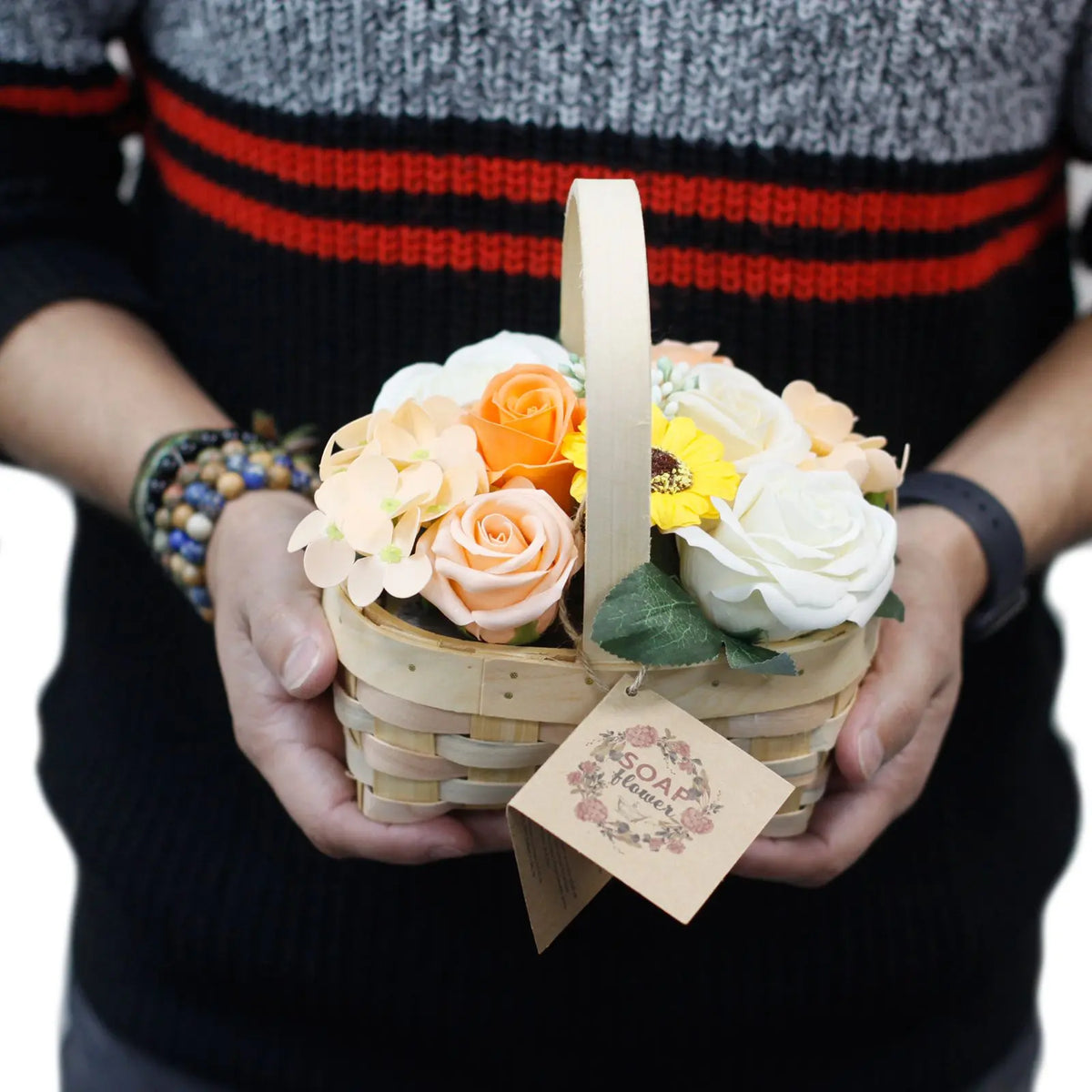 Persona sosteniendo una pequeña canasta de mimbre con flores de jabón en tonos naranja, amarillo y blanco.