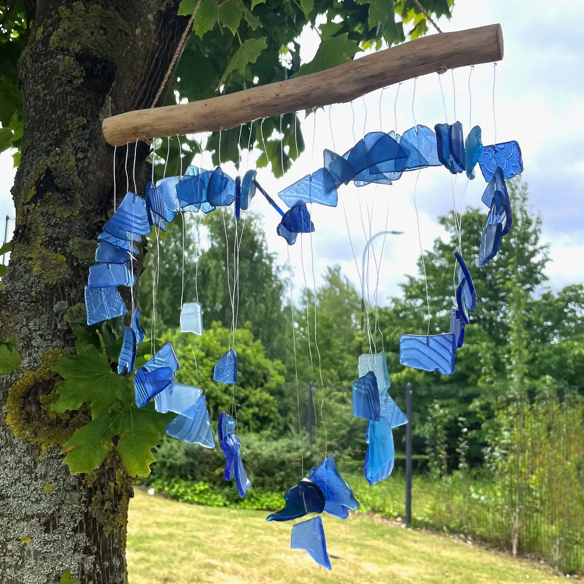 Carillón de piezas de vidrio reciclado azul y blanco, colgadas de una rama de madera natural en un jardín.