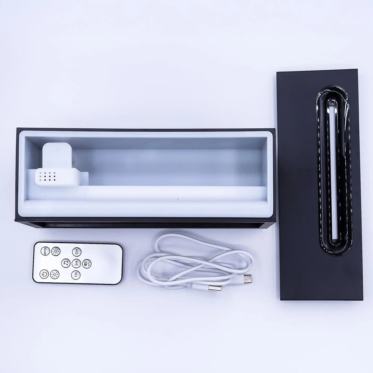 Difusor de aroma negro con tapa, cable USB y mando a distancia sobre fondo blanco. Vista superior de sus componentes.