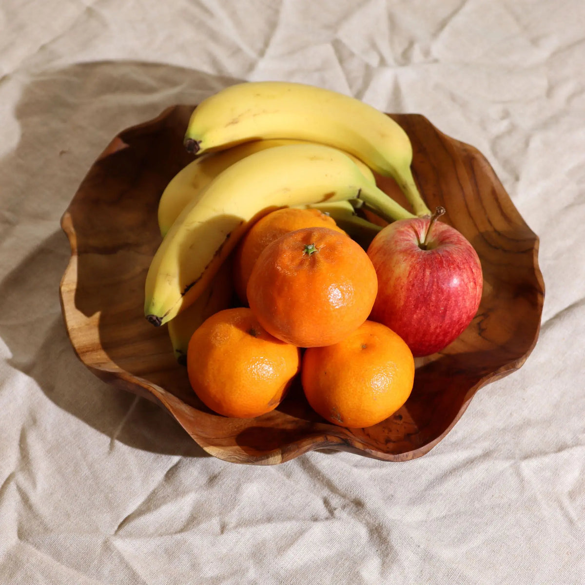 Cuenco artesanal de madera de teca con forma orgánica, lleno de plátanos, mandarinas y una manzana sobre tela clara.