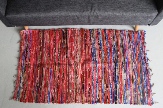 Alfombra de trapo rectangular (128x75cm) con rayas multicolores tejidas en tonos rojos, azules y neutros.
