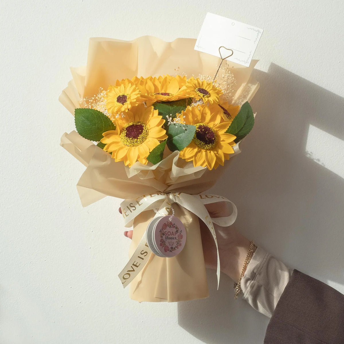 Ramo de flores de jabón con girasoles amarillos, hojas verdes y envoltura beige, decorado con una cinta blanca.