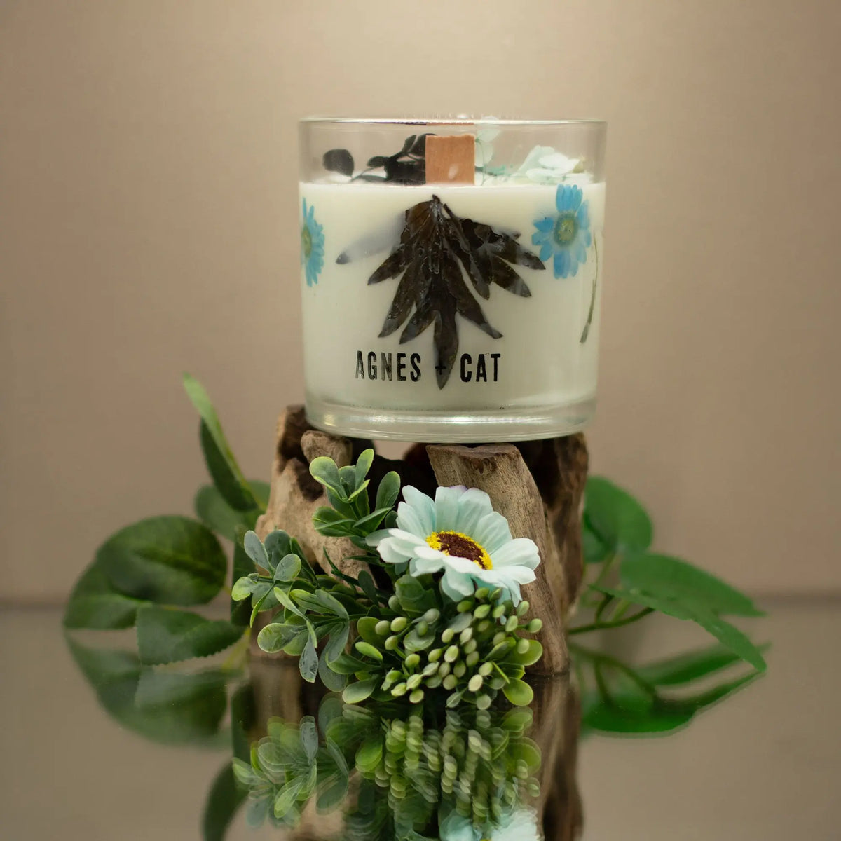 Vela aromática blanca con flores azules y madera, presentada en envase de vidrio, estilo natural y elegante.