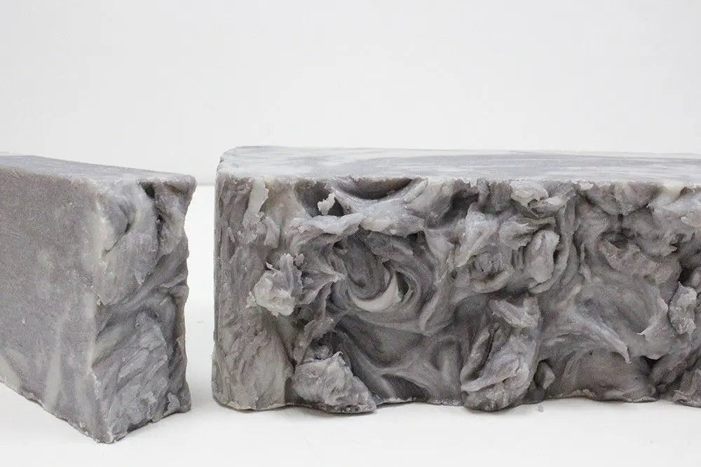 Barra de jabón artesanal de barro gris, con textura marmolada y rugosa, cortada en dos piezas sobre fondo blanco.