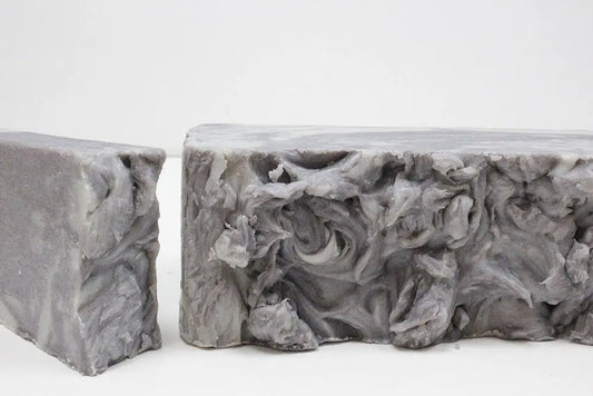 Barra de jabón artesanal de barro gris, con textura marmolada y rugosa, cortada en dos piezas sobre fondo blanco.