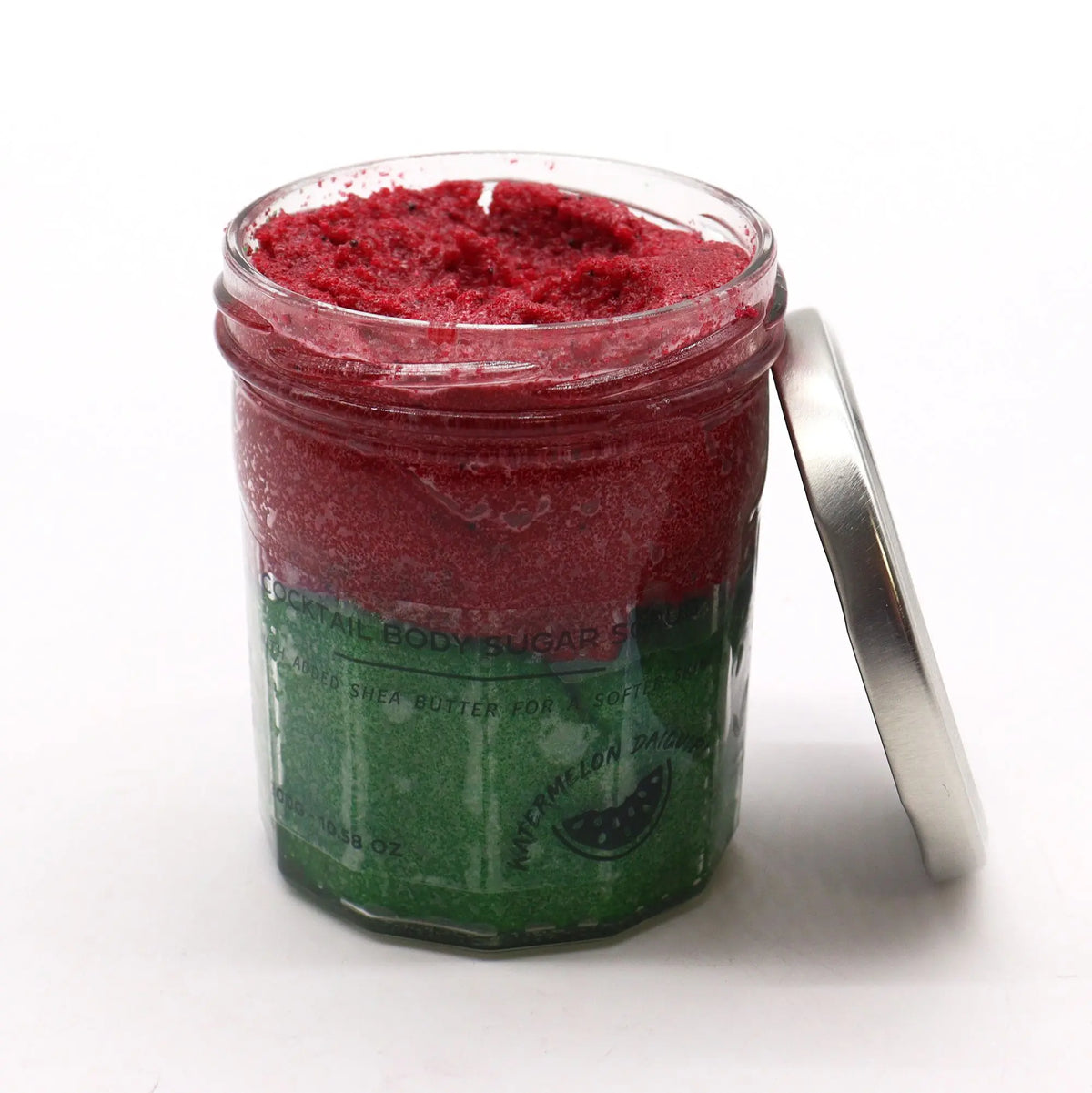 Frasco de vidrio con exfoliante de azúcar bicolor, rojo arriba y verde abajo, junto a una tapa metálica sobre fondo blanco.