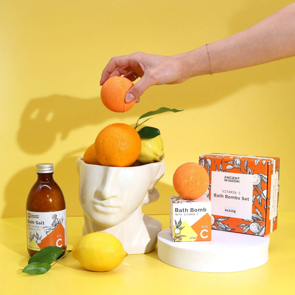 Set de 4 bombas de baño cítricas Ancient Wisdom. Incluye caja, sal de baño, frutas y escultura sobre fondo amarillo.