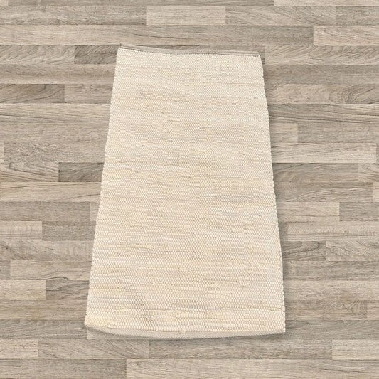 Alfombra rectangular de algodón color crema, tejida a mano con textura de cuentas, sobre suelo de madera.