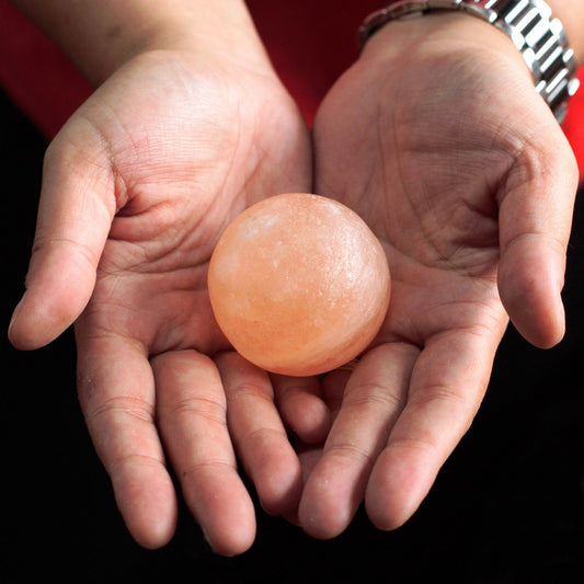 Dos manos sostienen una esfera de sal mineral del Himalaya color naranja claro, de textura suave y apariencia sólida.