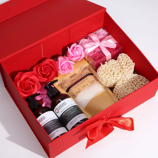 Caja de regalo roja, plegable, con lazo frontal, abierta y rellena con productos de belleza y flores decorativas.
