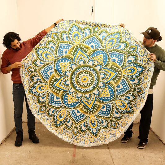 Dos personas sostienen un tapiz de algodón redondo con diseño de mandala en tonos amarillos y azules, con flecos.