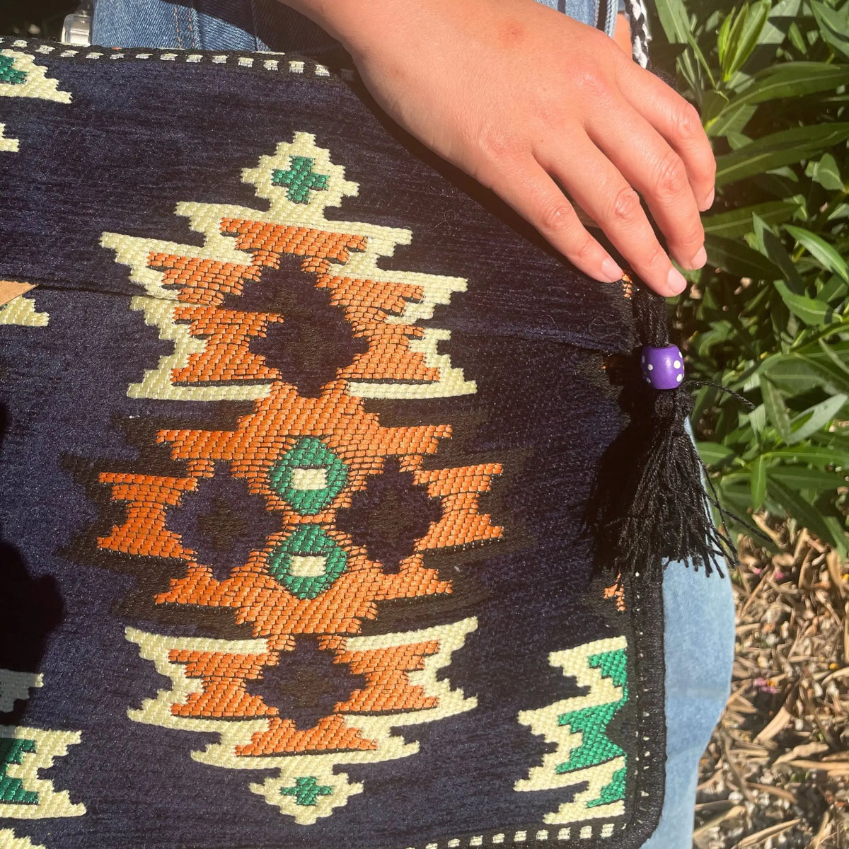 Bolso bandolera de tela azul marino con patrón kilim en tonos naranja y verde, detalle de borla y cuenta morada.
