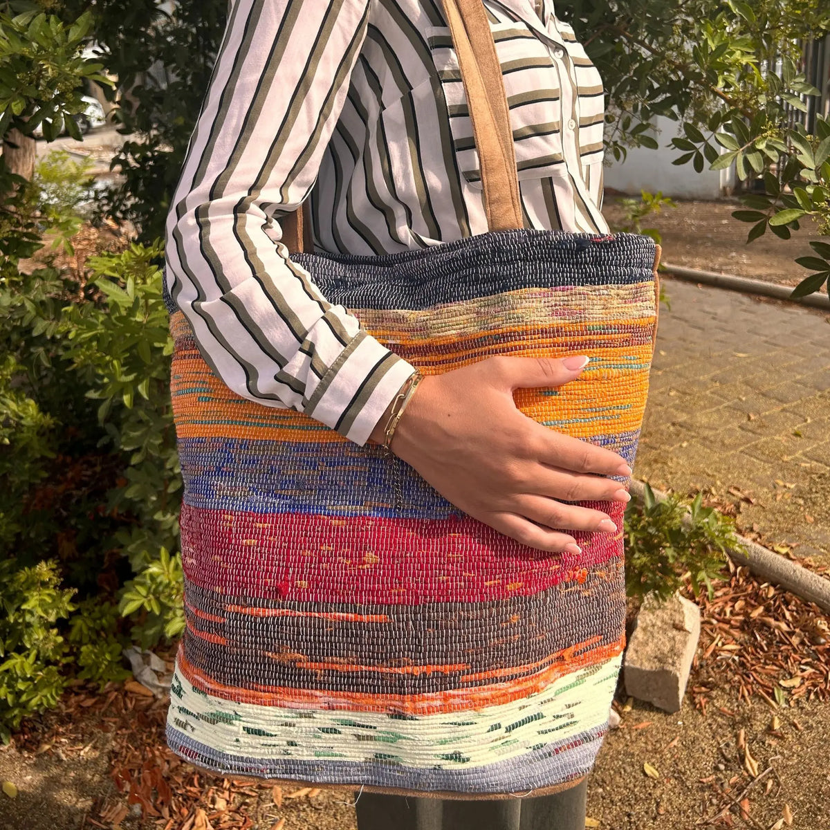Bolso tote grande de tejido reciclado con franjas coloridas, asas de cuero y diseño artesanal en exteriores.