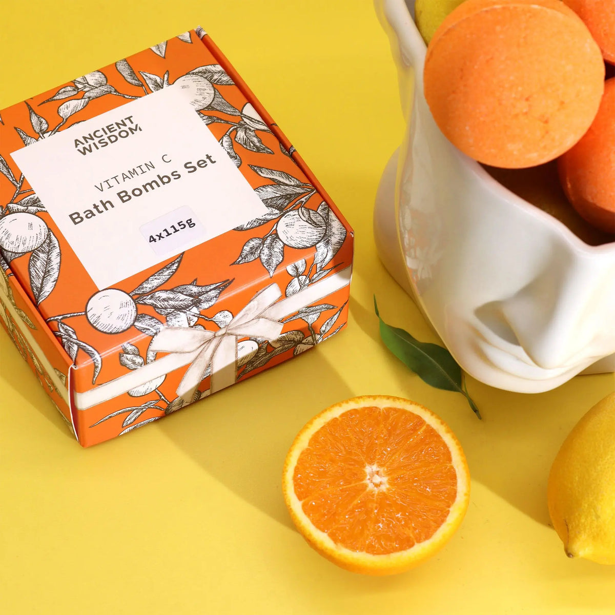 Set de 4 bombas de baño cítricas Ancient Wisdom en caja naranja con diseño de frutas, junto a naranjas y limones.