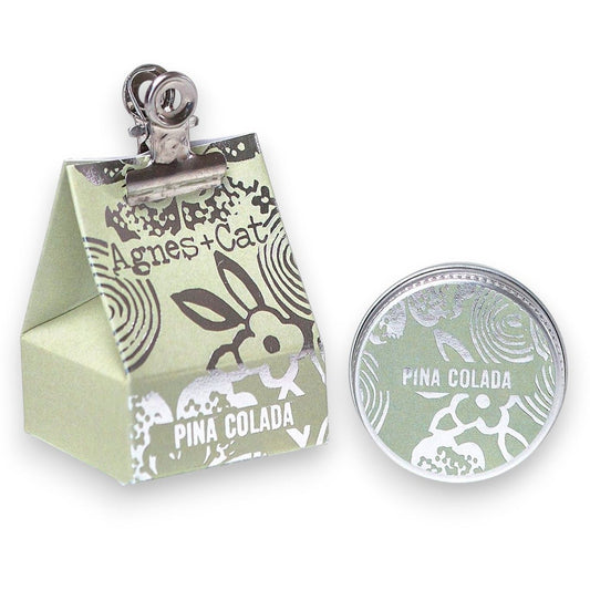 Bálsamo labial Piñacolada Ancient Wisdom en envase redondo y caja de papel verde con diseño floral y clip metálico.