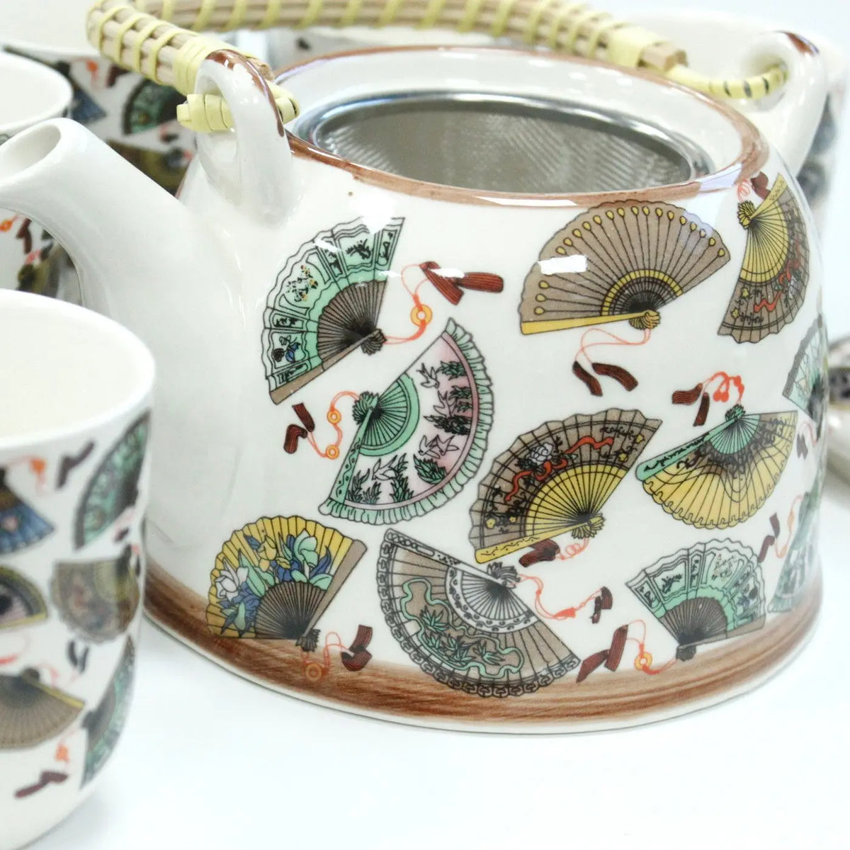 Tetera de cerámica blanca con diseño de abanicos, colador metálico y asa de bambú, acompañada de tazas a juego.