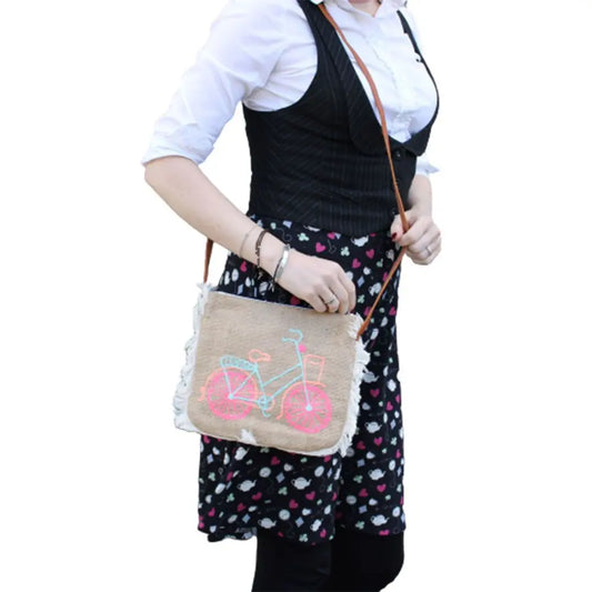 Bolso bandolera de yute con flecos laterales, decorado con un bordado de bicicleta en tonos azul y rosa neón.