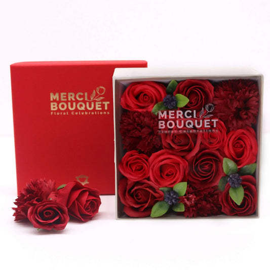 Caja cuadrada con flores de jabón rojas, incluyendo rosas y claveles, junto a su tapa roja. Estilo regalo elegante.