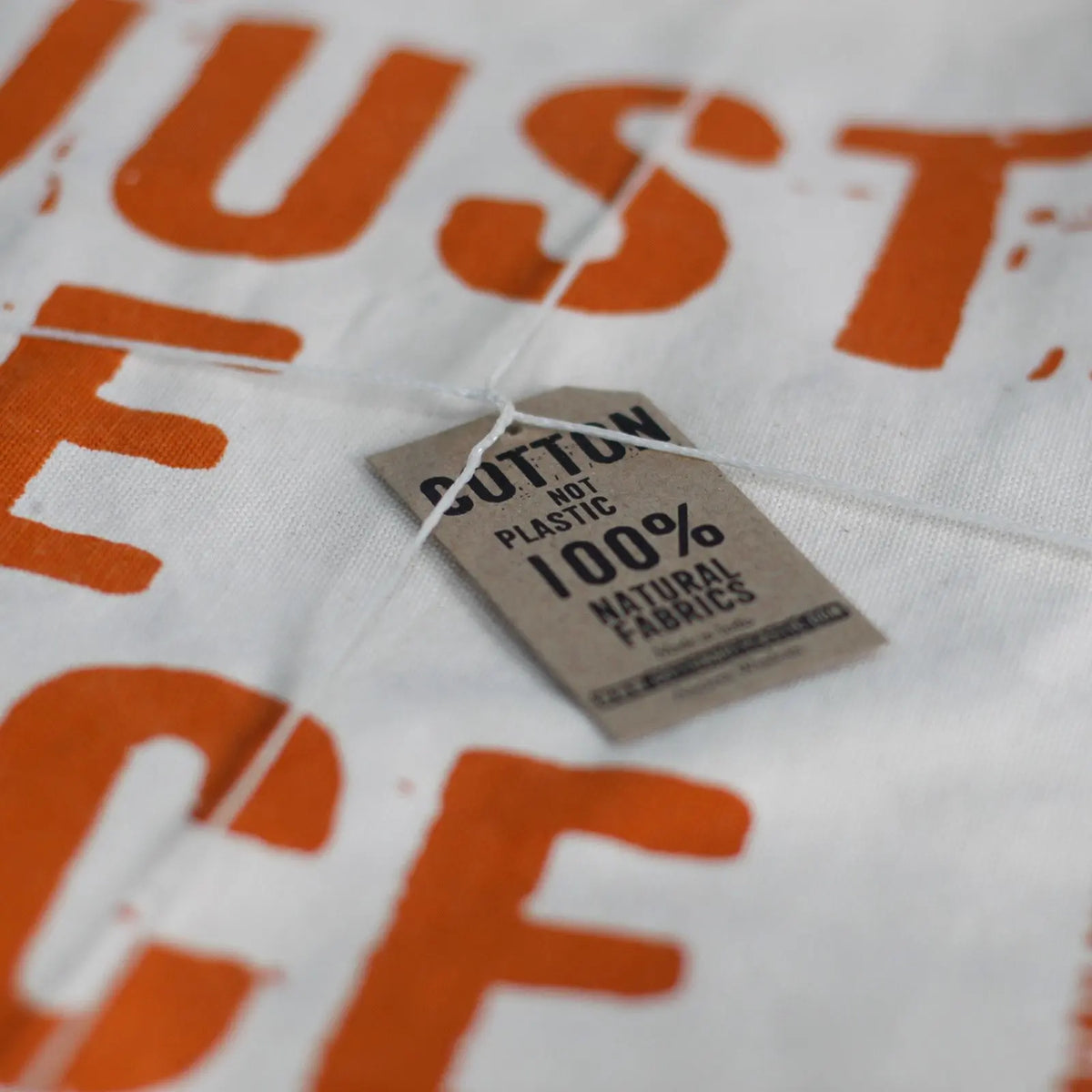 Bolsa de algodón crudo con letras naranjas y etiqueta de cartón con el texto "100% natural fabrics".