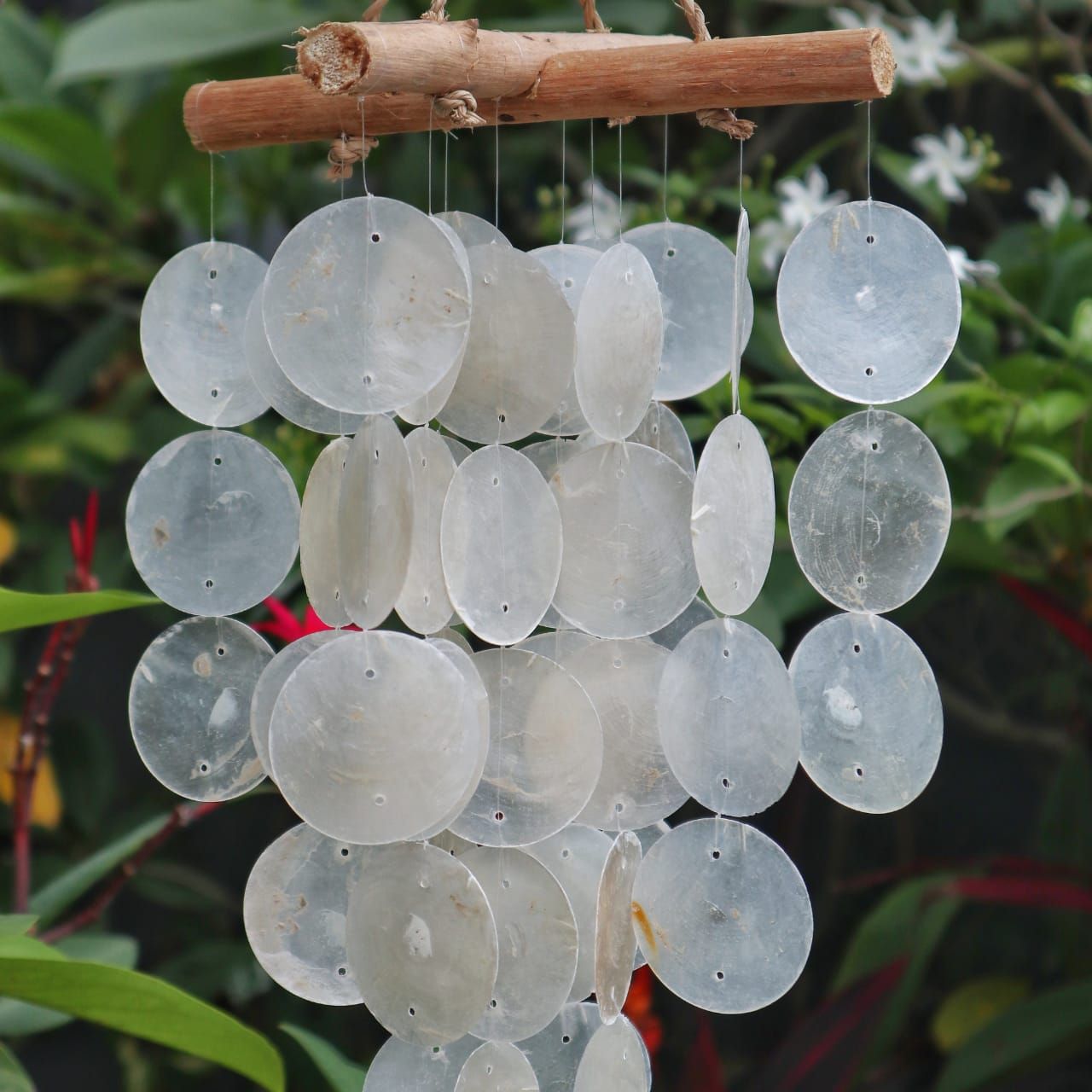 Carillón decorativo con discos de nácar colgando de ramas de madera natural en un entorno de jardín con vegetación