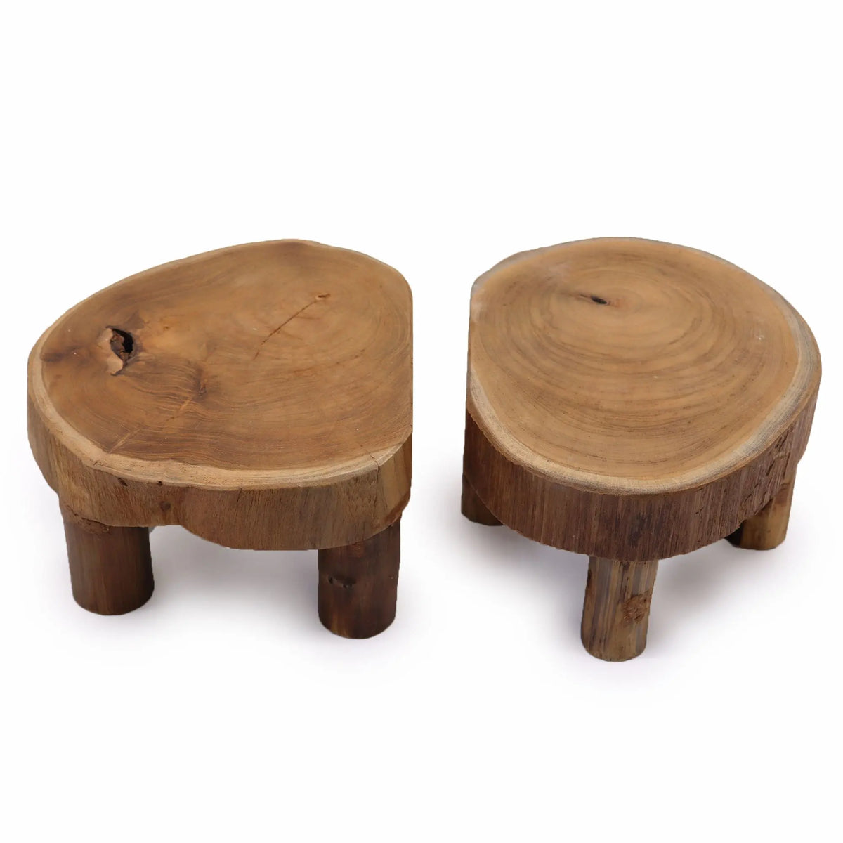 Dos soportes bajos de madera de teca natural con superficie redonda irregular y tres patas, sobre fondo blanco.
