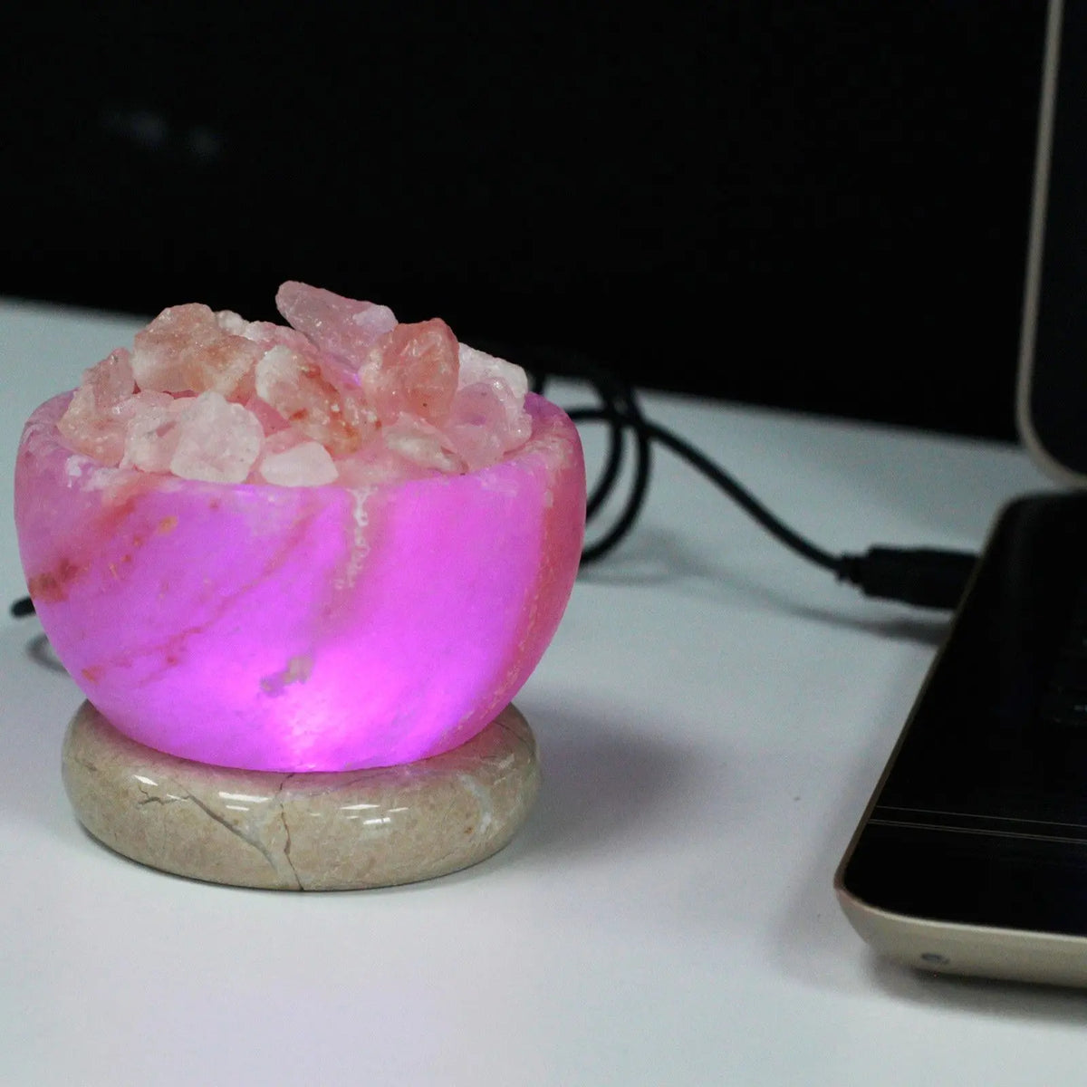 Lámpara de sal tipo cuenco con cristales rosados iluminados en luz LED rosa sobre base circular, conectada por USB.