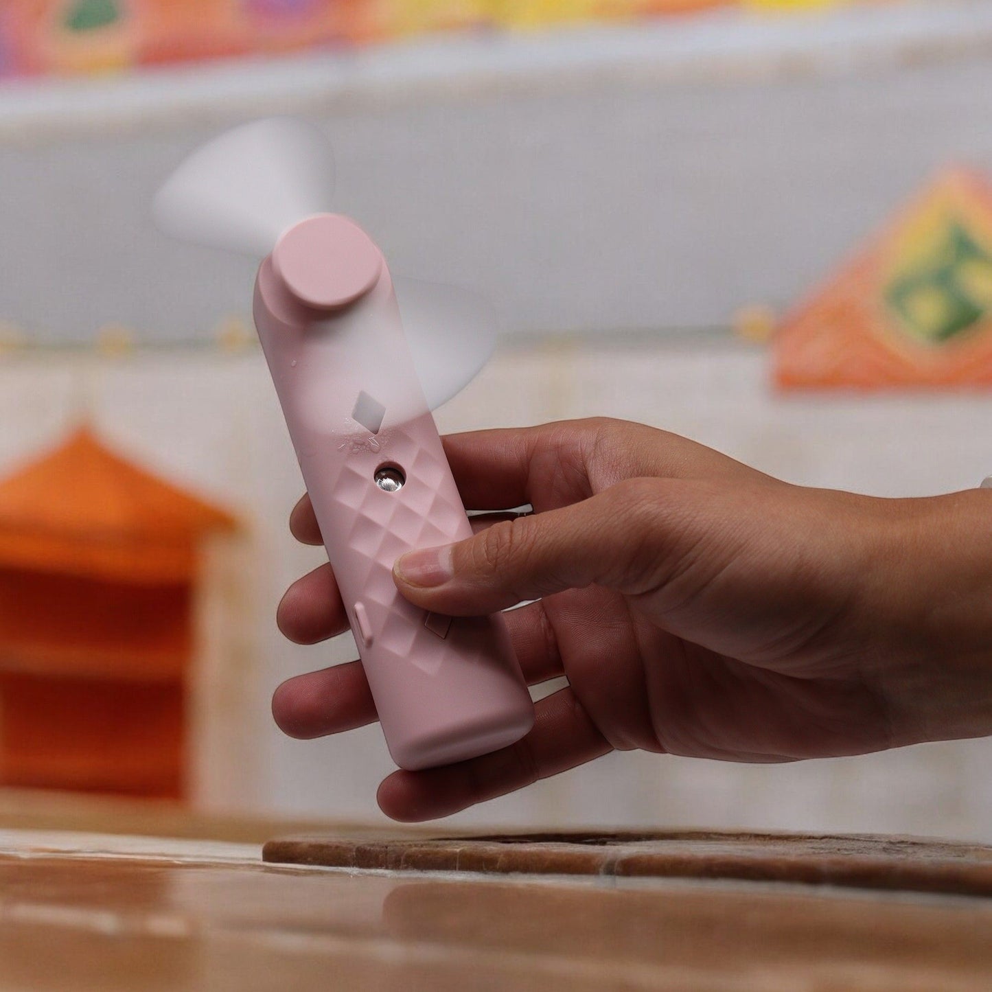 Mano sosteniendo ventilador pulverizador facial rosa de plástico, portátil, con textura romboidal y aspas en movimiento.